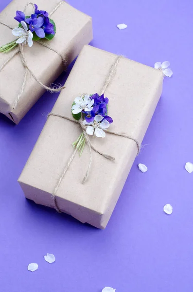 Hermosas cajas de regalo envueltas en papel artesanal marrón simple decorado con flores vivas de violeta sobre un fondo violeta . —  Fotos de Stock