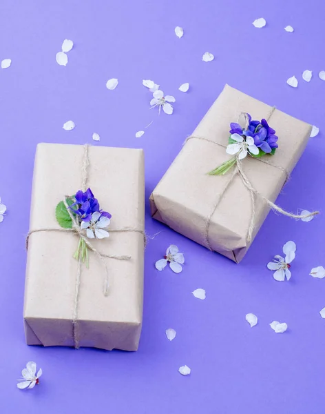 Belles boîtes-cadeaux enveloppées dans du papier artisanal marron simple décoré de fleurs vivantes de violette sur fond violet . — Photo
