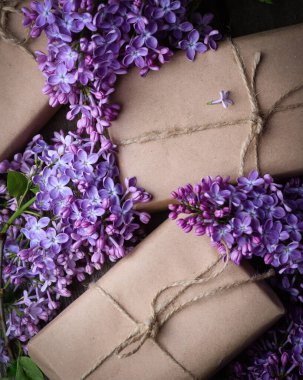 El yapımı, kahverengi kağıtlarla sarılı, eski ahşap bir yüzeyde taze mor leylak çiçekleriyle süslenmiş hediye kutuları. Bahar tatili geçmişi.