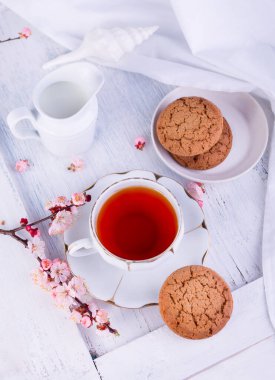 Beyaz porselen çay, süt sürahisi ve taze pişmiş yulaf kurabiyesi. İngiliz kahvaltısı, içecek, ikram ve beyaz masa örtüsü ile natürmort..
