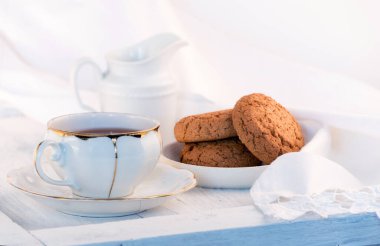 White porcelain cup of tea, milk jug, and fresh-baked oat cookies. English breakfast still-life with drink and treats and white tablecloth. clipart