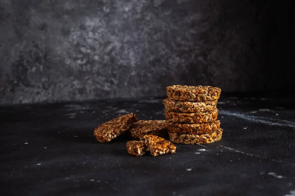 Caramel peanut brittle from sunflower seeds and sesame seeds and fitness cookies with flax seeds on dark black concrete background.