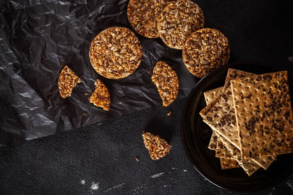 Caramel peanut brittle from sunflower seeds and sesame seeds and fitness cookies with flax seeds on dark black concrete background.