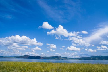 Çiçekli bozkır ve Sibirya nehri üzerinde bulutlarla kaplı mavi bir gökyüzü. Arka planda dağlar. Krasnoyarsk bölgesi