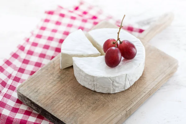 ぶどうと赤の市松模様のテーブル クロスで木の板にチーズの部分と柔らかいチーズ ホイール — ストック写真