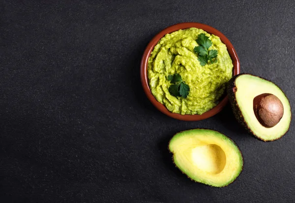 Salsa di guacamole in ciotola di argilla con metà avocado tagliato su bac scuro — Foto Stock