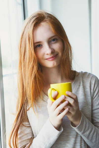 candid portrait of cheerful young smiling red hair woman holding yellow coffee cup, enjoying morning coffee and new day, waking up