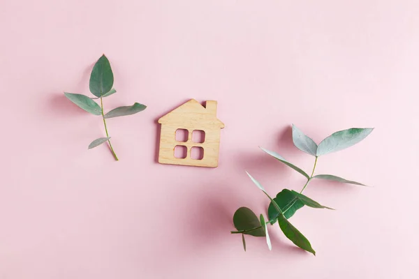 little wooden ecological house and eucalyptus on pastel pink background, top view, flat lay, symbol  of shelter, accommodation, sweet home concept