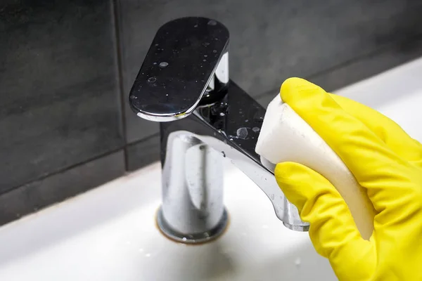 Рука в жёлтой резиновой перчатке вытирает грязный хромированный металлический кран белой губкой из меламина. Уборка в современной ванной комнате. Выборочный фокус. Вид крупным планом — стоковое фото