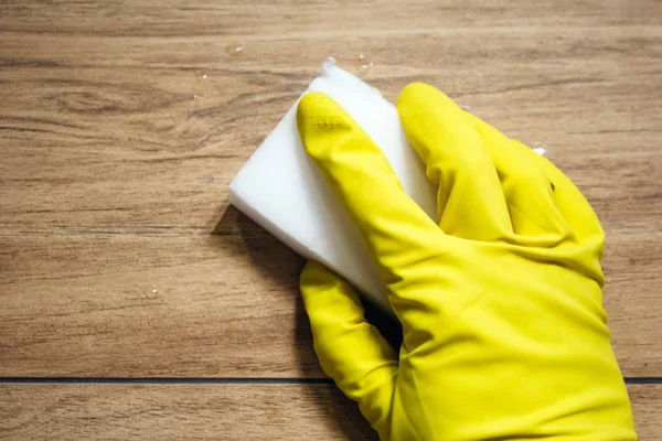 Uma mão em uma luva de borracha amarela limpa a telha molhada imitando uma superfície de madeira com uma esponja de melamina branca. Limpeza em uma casa de banho moderna. Foco seletivo — Fotografia de Stock