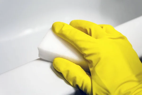 Uma mão em uma luva de borracha amarela limpa a superfície de um banho branco com uma esponja de melamina. Limpeza em uma casa de banho moderna. Foco seletivo. Vista de perto — Fotografia de Stock