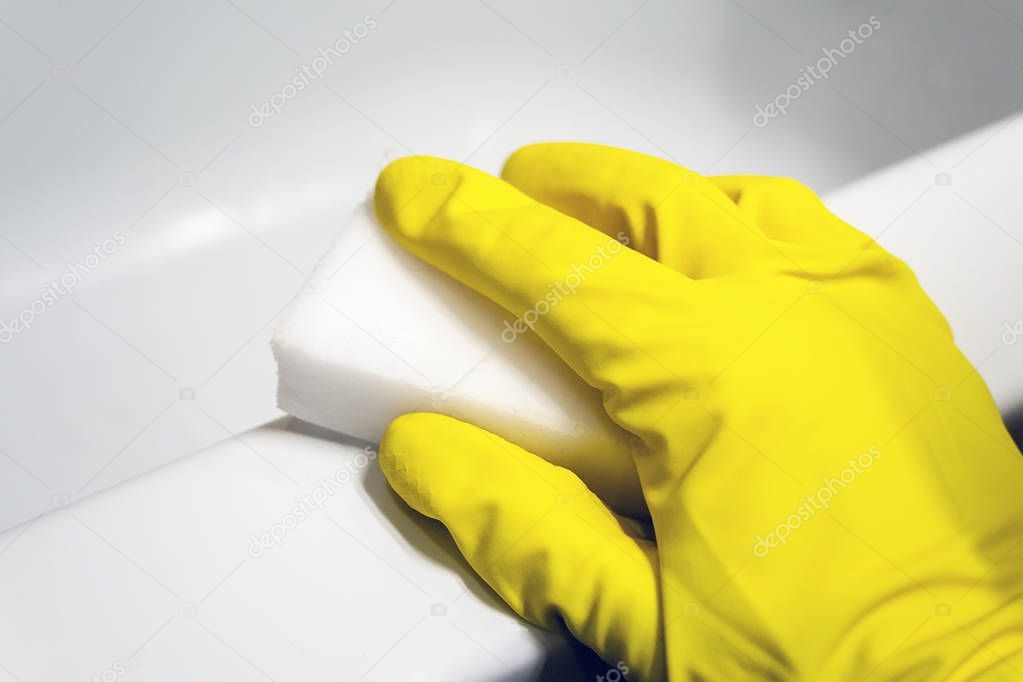 A hand in a yellow rubber glove wipes the surface of a white bath with a melamine sponge. Cleaning in a modern bathroom. Selective focus. Closeup view