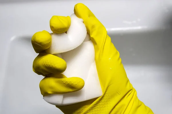 A hand in a yellow rubber glove squeezes a wet white melamine sponge against a white bathroom. Selective focus. Closeup view — Stock Photo, Image