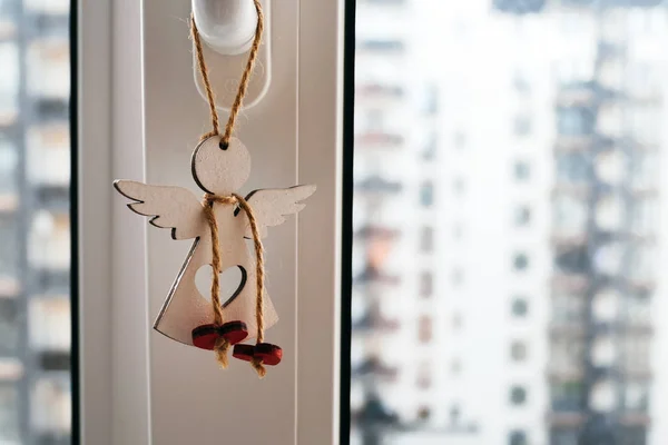 Деревянный ангел с отверстием в форме сердца, висящим на восковой веревке на оконной ручке на размытом фоне соседних домов. Защитное обаяние с красными сердцами на кружеве вокруг шеи — стоковое фото