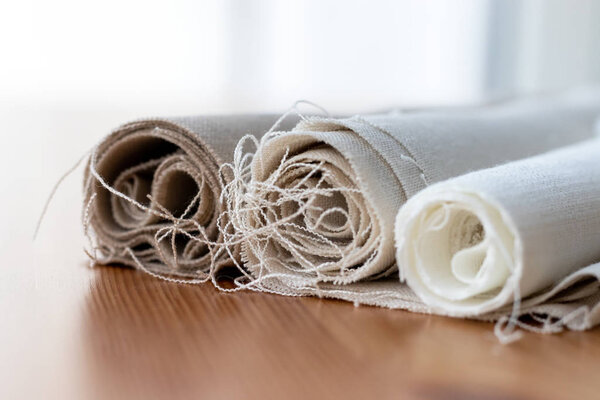 Three rolls of natural linen fabric in different colors with protruding threads on a wooden table. Selective focus. Closeup view. Blurred background
