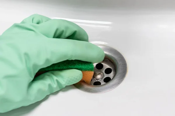 Una mano in un guanto di gomma verde menta con una spugna arancione strofina il bagno bianco sporco bagnato o il lavandino vicino al foro di scarico. Pulizia in un bagno moderno. Concentrazione selettiva. Vista da vicino — Foto Stock