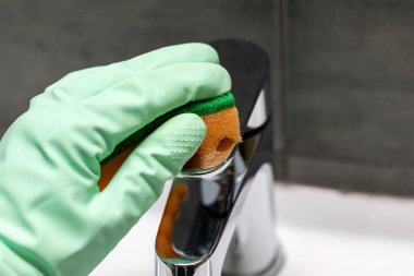 A hand in a mint green rubber glove wipes a chrome metal faucet with an orange sponge. Cleaning in a modern bathroom. Selective focus. Closeup view