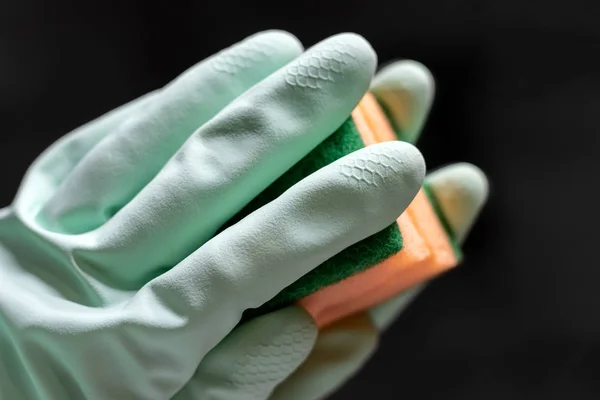 Рука в мятной зеленой резиновой перчатке вытирает зеркало в темной комнате оранжевой губкой. Вид крупным планом. Селективный фокус — стоковое фото
