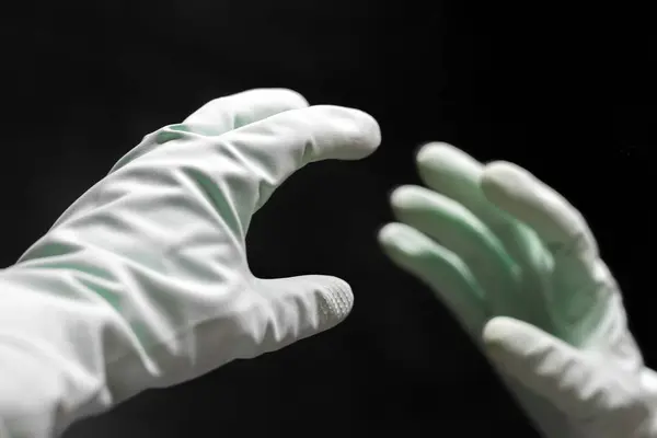 A hand in a mint green rubber glove reaches for its reflection in the mirror in the dark black room. Selective focus. Closeup view — 스톡 사진