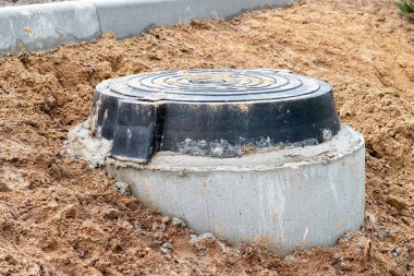 Beton bir üsteki siyah dökme demir kanalizasyon kapağı. Yeni bir yerleşim yerinin inşaat sahasındaki kanalizasyon kuyusuna ulaşmak için nemli bir zeminde bulunuyor.