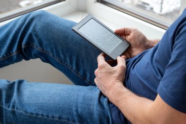 Mavi tişörtlü ve kot pantolonlu bir adam elinde elektronik bir kitap tutuyor. Evde, pencerenin yanında kitap okuyordum. Seçici odak