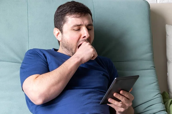 Adam e-kitap okurken esniyor. Mavi tişörtlü, dinlenen bir erkek bir elinde elektronik kitapla yeşil bir koltukta oturuyor, esniyor, ağzını ikinci eliyle kapatıyor.