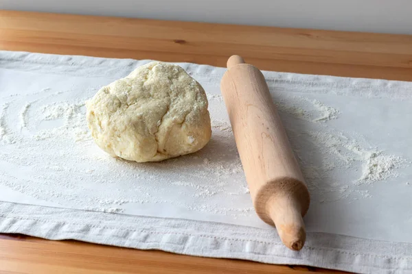 Raw curd dough with bits of cottage cheese, wooden rolling pin and sprinkled flour on baking paper on a linen tablecloth on a wooden table. The process of making curd bagels or croissants