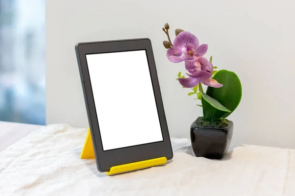 Mutfak masasının üzerinde pencere kenarında keten masa örtüsü olan yapay pembe bir orkidenin yanında sarı bir ekranda boş bir maket ekranı olan modern gri elektronik kitap veya tablet.