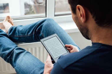 Mavi kotlu bir adamın, pencerenin kenarında, parlak, modern bir apartmanın pencere eşiğinde çıplak ayakla duran elektronik bir kitap okuduğu omuz manzarası.