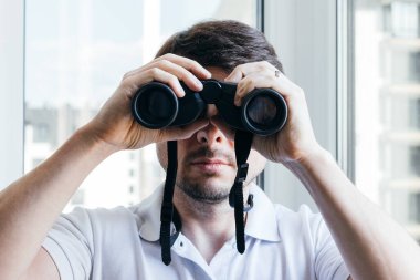 Karantina sırasında çevreyi ya da doğayı gözlemlemek için komşu evi gören yakışıklı bir adam pencerenin yanındaki kameraya dürbünle bakıyor. Seçici odaklanma. Yakın görünüm