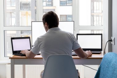 Beyaz tişörtlü bir adam iş yerinde iki dizüstü bilgisayar ve pencerenin yanında bir monitörle oturuyor. Karantinadaki evden uzak bir çalışma. Arka plan