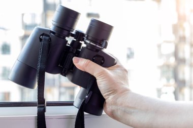 Siyah bir dürbünle komşu evi, çevreyi ya da doğayı gözetleyen bir pencerenin bulanık arka planına karşı el ele tutuşmak. Seçici odaklanma. Yakın görünüm