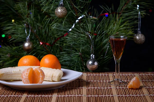 Bir tabakta mandalinalı muz ve masanın yüzeyinde bir bardak alkol Noel ağacının arka planına karşı..