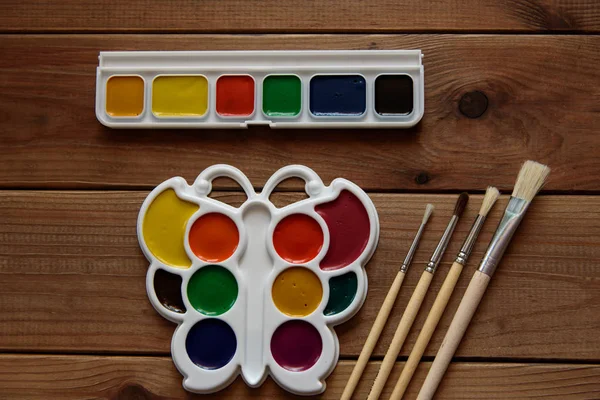 Beyaz plastik bir palette renkli kuru suluboya tabloları ve ahşap bir masada fırçalar..