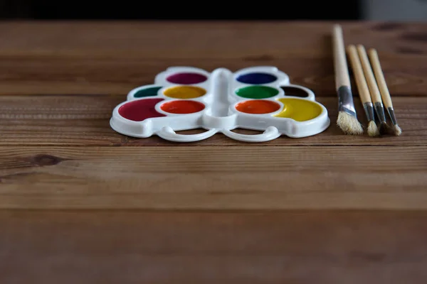 Beyaz plastik bir palette renkli kuru suluboya tabloları ve ahşap bir masada fırçalar..