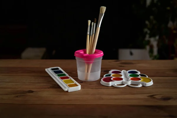 Beyaz plastik bir palette renkli kuru suluboya tabloları ve ahşap bir masada fırçalar..