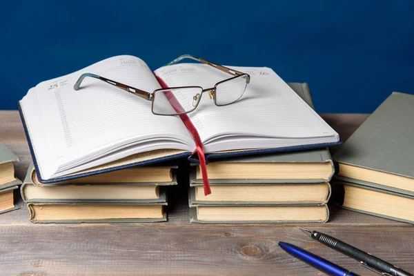 Masanın üzerinde açık bir defter, bir büyüteç, bir kalem ve kitaplar var.