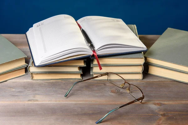 Masanın üzerinde açık bir defter, bir büyüteç, bir kalem ve kitaplar var.