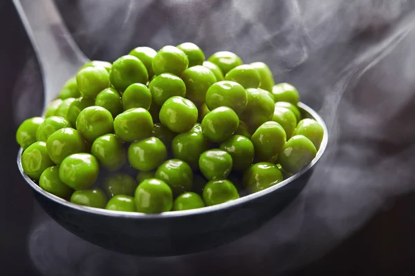 Groene erwten in een lepel met stoom tegen een donkere achtergrond. Sluiten. — Stockfoto