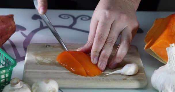 A nő narancspaprikát szeletel egy fa deszkán a konyhában. A női kezek elkészítik a salátát paprikával. Egészséges táplálkozás koncepciója — Stock videók