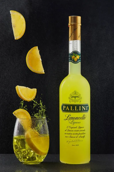 Láhev italského alkoholického likéru Limoncello, sklenici s ledem a nápoj, do kterého na černé pozadí dopadají plátky citronu. — Stock fotografie