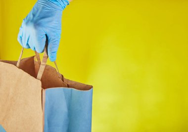 Mavi eldivenli teslimatçı işvereni sarı stüdyo arka planında izole edilmiş yiyecek dolu el işi kağıt torba tutuyor. Servis karantina salgını Coronavirus virüsü konsepti 2019-ncov. Boşluğu kopyala.