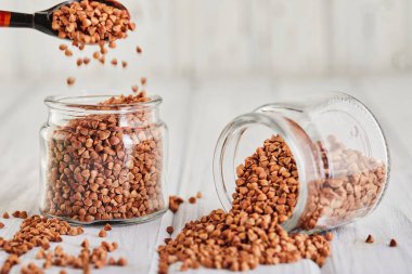 Dağıtılmış taze karabuğday tarlaları beyaz ahşap arka planda bir kaşıktan cam bir kavanoza serpilmiş. Kopya Boşluğu.