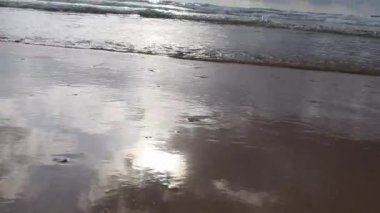 4K Video Manzarası, günbatımında kumlu bir sahile vuran mavi deniz suyu dalgalarının geniş bir görüntüsü. Güneş parıldıyor, deniz yüzeyini gözetliyor..