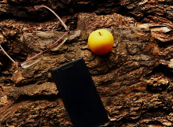 Yellow apple, glasses and cellphone flat lay on tree bark