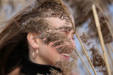 Dışarıdaki kameraya poz veren güzel bir genç kadın. Hızlı bir geçmiş. Açık havada. Kapatın. Kuru sazlığın ve mavi gölün yakınında güneşli bir günde. Kahverengi ceketli, uzun saçlı, güzel bir sarışın.