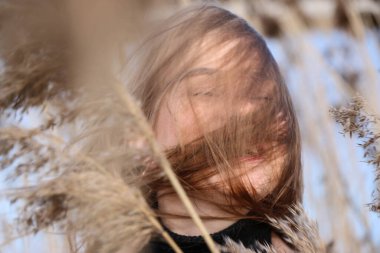Dışarıdaki kameraya poz veren güzel bir genç kadın. Hızlı bir geçmiş. Açık havada. Kapatın. Kuru sazlığın ve mavi gölün yakınında güneşli bir günde. Kahverengi ceketli, uzun saçlı, güzel bir sarışın.