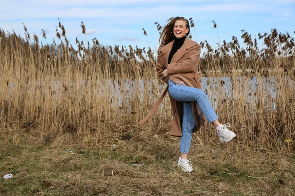 Outdoors lifestyle fashion portrait of happy stunning blonde girl. Beautiful smile. Long light hair. Wearing stylish coat. Joyful and cheerful woman. walk on a natural landscape, near a dry reed and a lake on a sunny day.