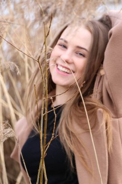 Dışarıdaki kameraya poz veren güzel bir genç kadın. Hızlı bir geçmiş. Açık havada. Kapatın. Kuru sazlığın ve mavi gölün yakınında güneşli bir günde. Kahverengi ceketli, uzun saçlı, güzel bir sarışın.