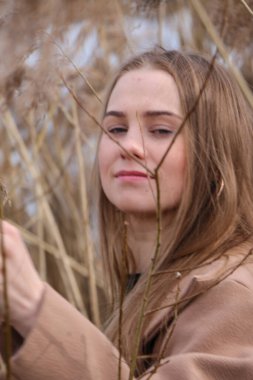 Dışarıdaki kameraya poz veren güzel bir genç kadın. Hızlı bir geçmiş. Açık havada. Kapatın. Kuru sazlığın ve mavi gölün yakınında güneşli bir günde. Kahverengi ceketli, uzun saçlı, güzel bir sarışın.